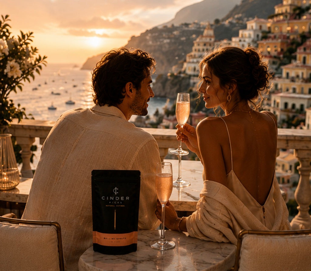 Couple enjoying a peach bellini at sunset with a scenic background, Cinder Picks packaging in the foreground.