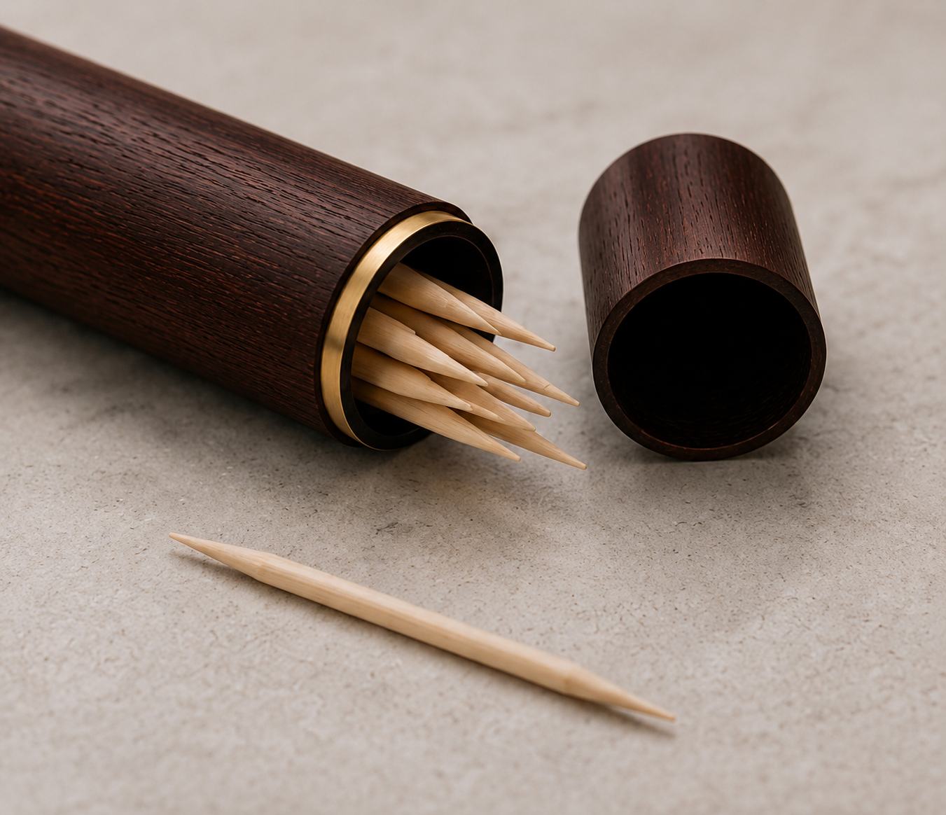 A dark wood toothpick holder with a gold-tone metal band, shown open with several birchwood toothpicks inside and one toothpick lying in front, on a neutral textured surface.