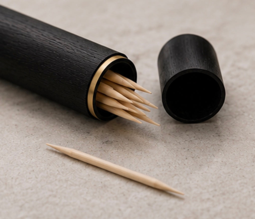 A black  Cinder Picks flavoured toothpick holder with a gold-toned rim, shown open with several birchwood toothpicks inside and one loose toothpick on a neutral textured surface.