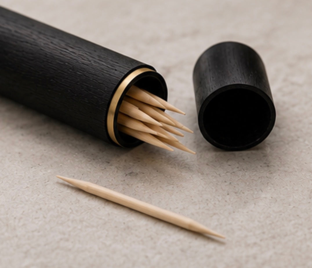 A black  Cinder Picks flavoured toothpick holder with a gold-toned rim, shown open with several birchwood toothpicks inside and one loose toothpick on a neutral textured surface.