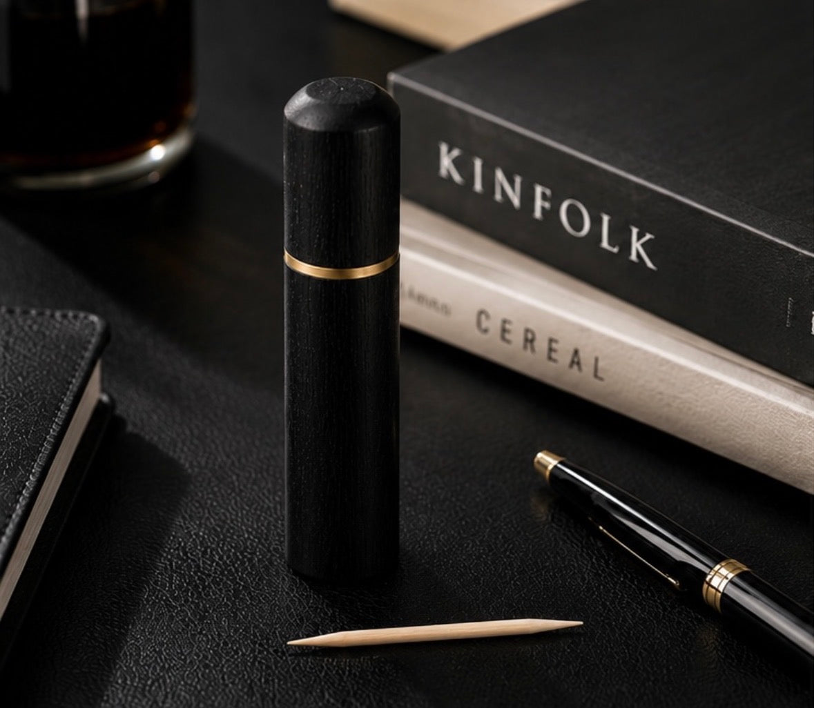 A black cylindrical pen or accessory with a thin gold band stands upright on a dark tabletop, surrounded by a black pen, a Cinder Picks toothpick, stacked books including one labelled KINFOLK, and part of a glass in a moody office-style setting.