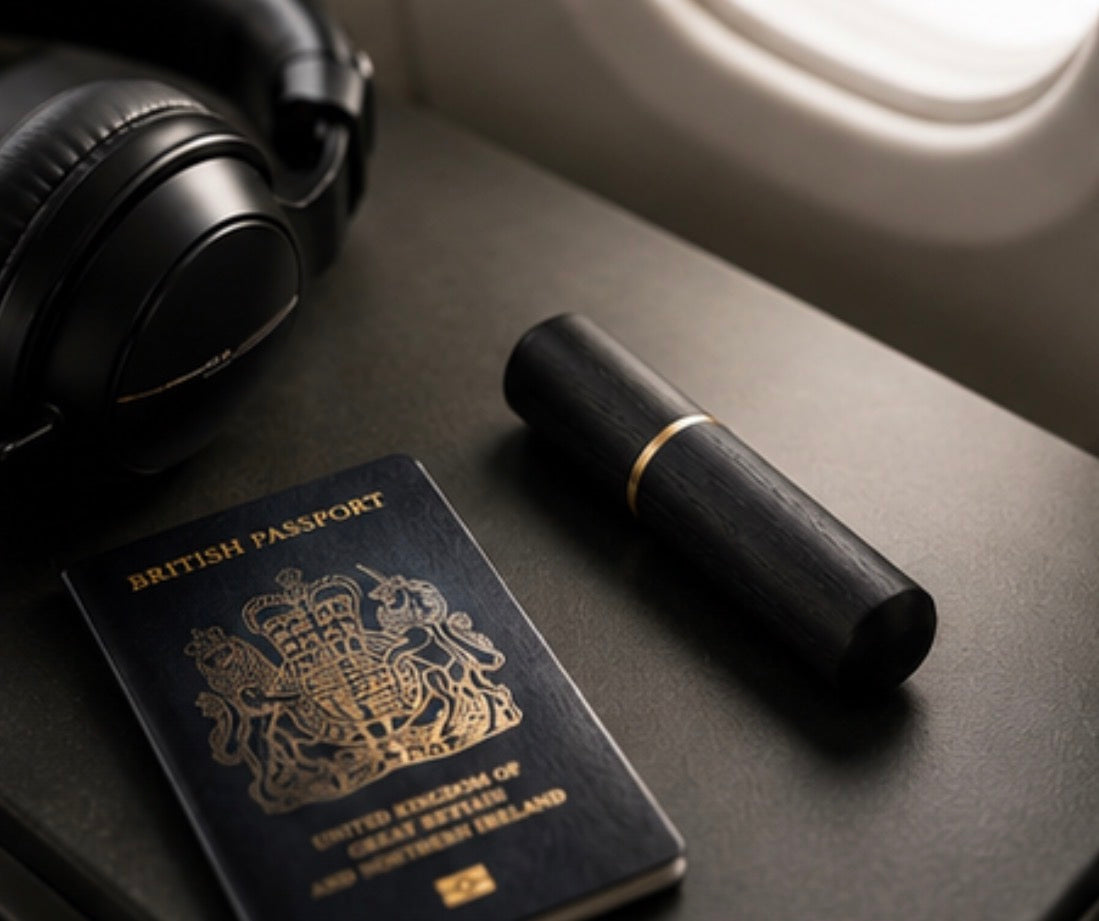 A black cylindrical  Cinder Picks flavoured toothpick holder with a gold band lies on a plane tray table beside a British passport, black over-ear headphones, and an airplane window.