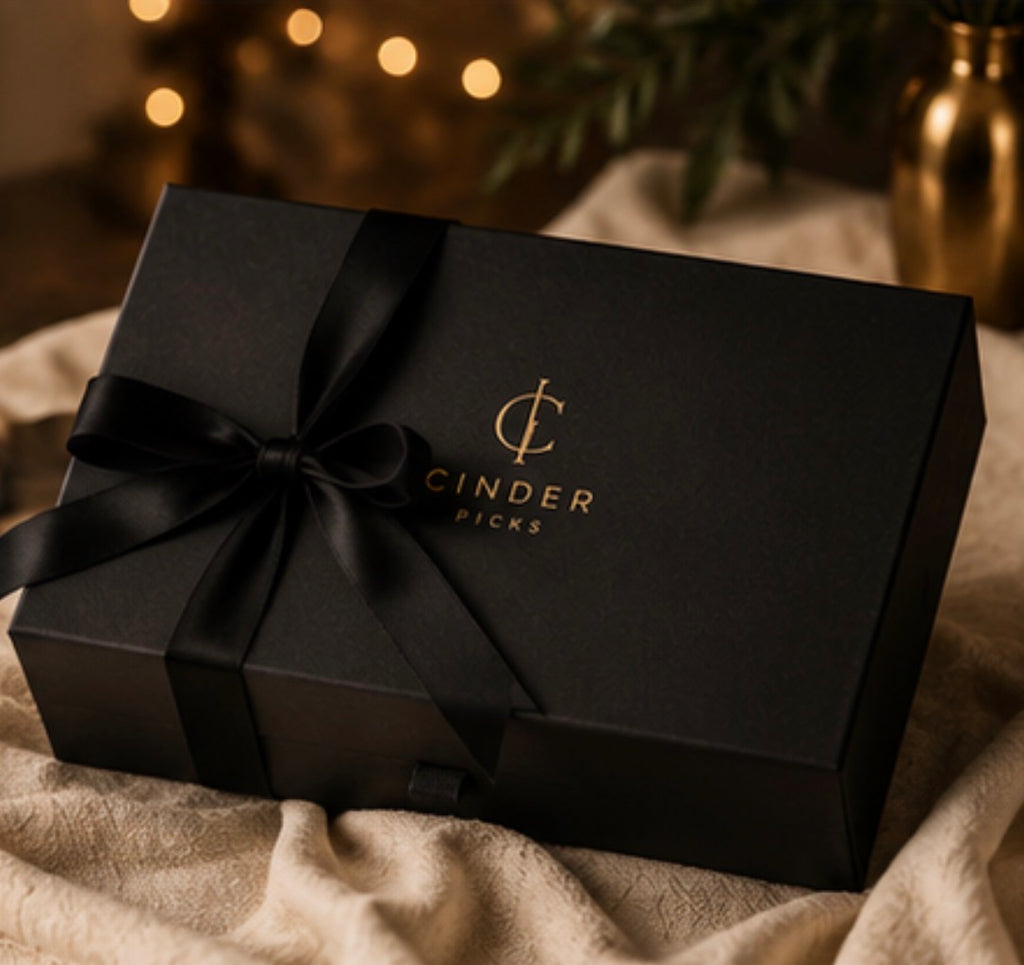 Black gift box with a bow and 'Cinder Picks' logo on a textured surface with blurred lights in the background.