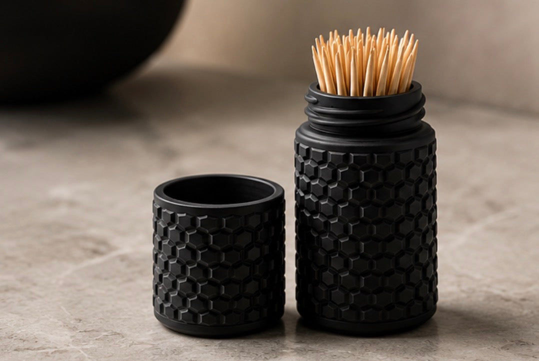 A black textured toothpick holder set with a honeycomb pattern, shown with the lid off and toothpicks inserted, on a neutral stone surface.