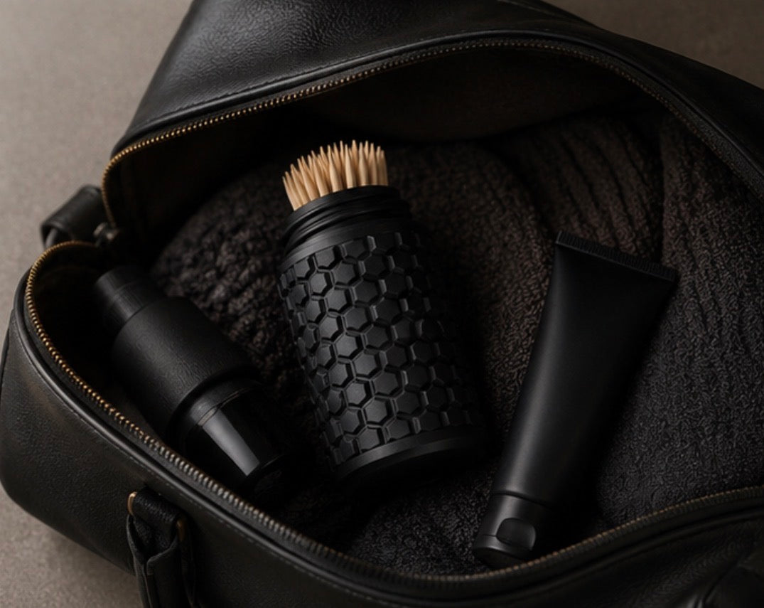 A black leather zippered travel or toiletry bag opened to reveal several black grooming or personal care items inside, including a textured cylindrical toothpick holder, a black bottle, and a black tube, set against a neutral grey background.