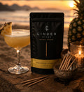 Cinder Picks Pineapple Daquiri packaging with a cocktail and pineapple on a beach at sunset.