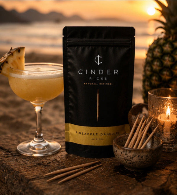 Cinder Picks Pineapple Daquiri packaging with a cocktail and pineapple on a beach at sunset.