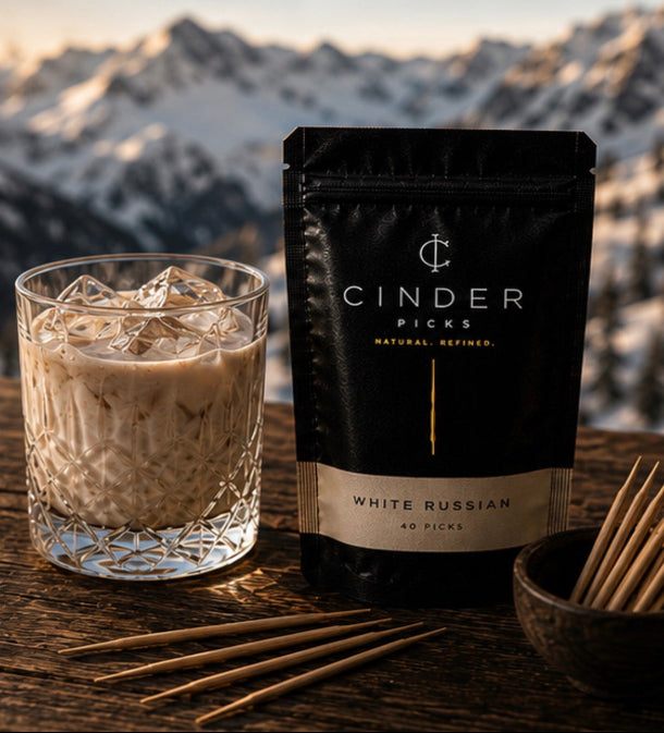 Glass of a White Russian cocktail with ice and a package of Cinder Picks against a mountainous background.