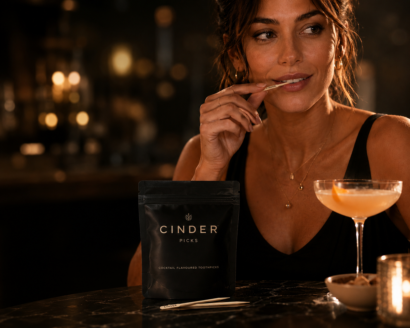 A woman in a black dress sits at a dimly lit bar, holding a Cinder Pick near her mouth. In front of her is a black pouch labelled CINDER PICKS and a cocktail glass with an orange drink.